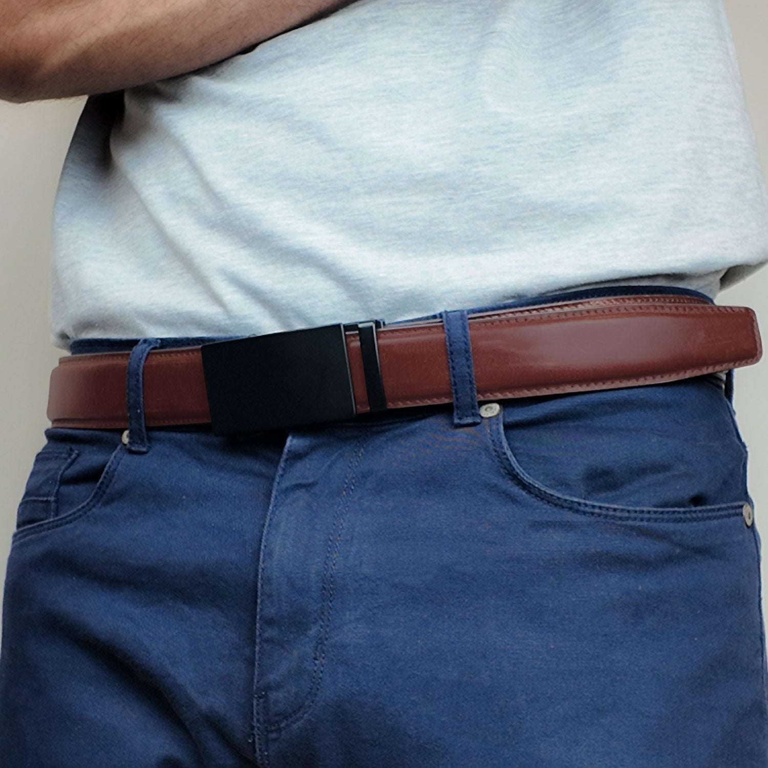 A man wearing blue jeans and a light blue shirt highlights a tan brown leather ratchet belt with a shiny black rectangular buckle, offering a sleek and functional design.